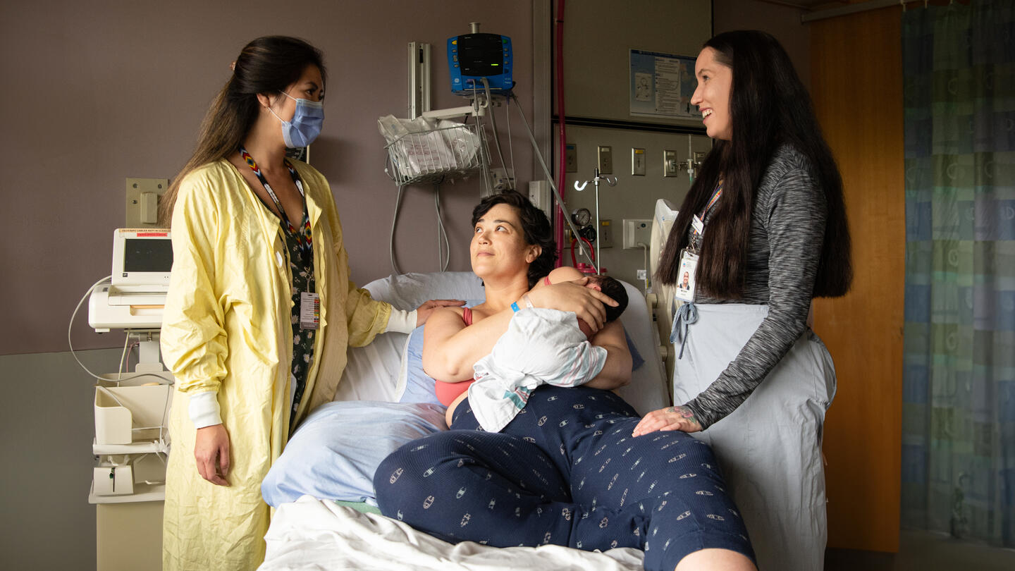 Two women stand next to a woman in a hospital bed holding a baby