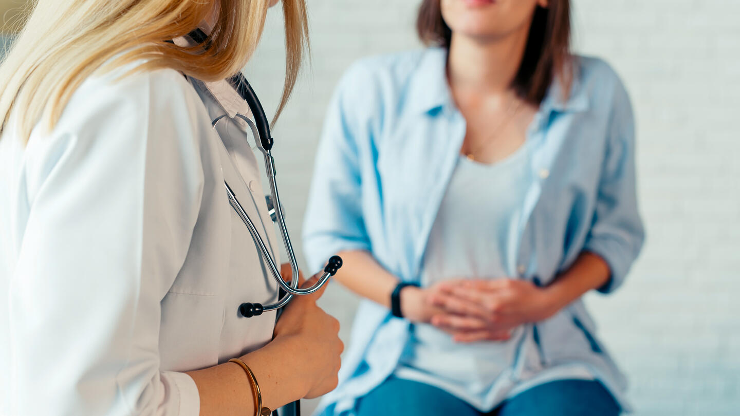 A doctor stands in the foreground looking at a pregnant woman with her hands on her stomach