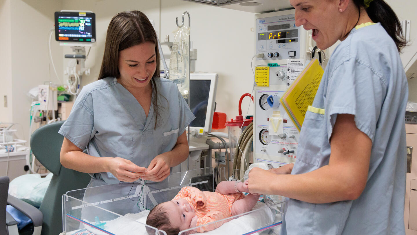 Two women in scrubs stand looking at a baby in a bassinet