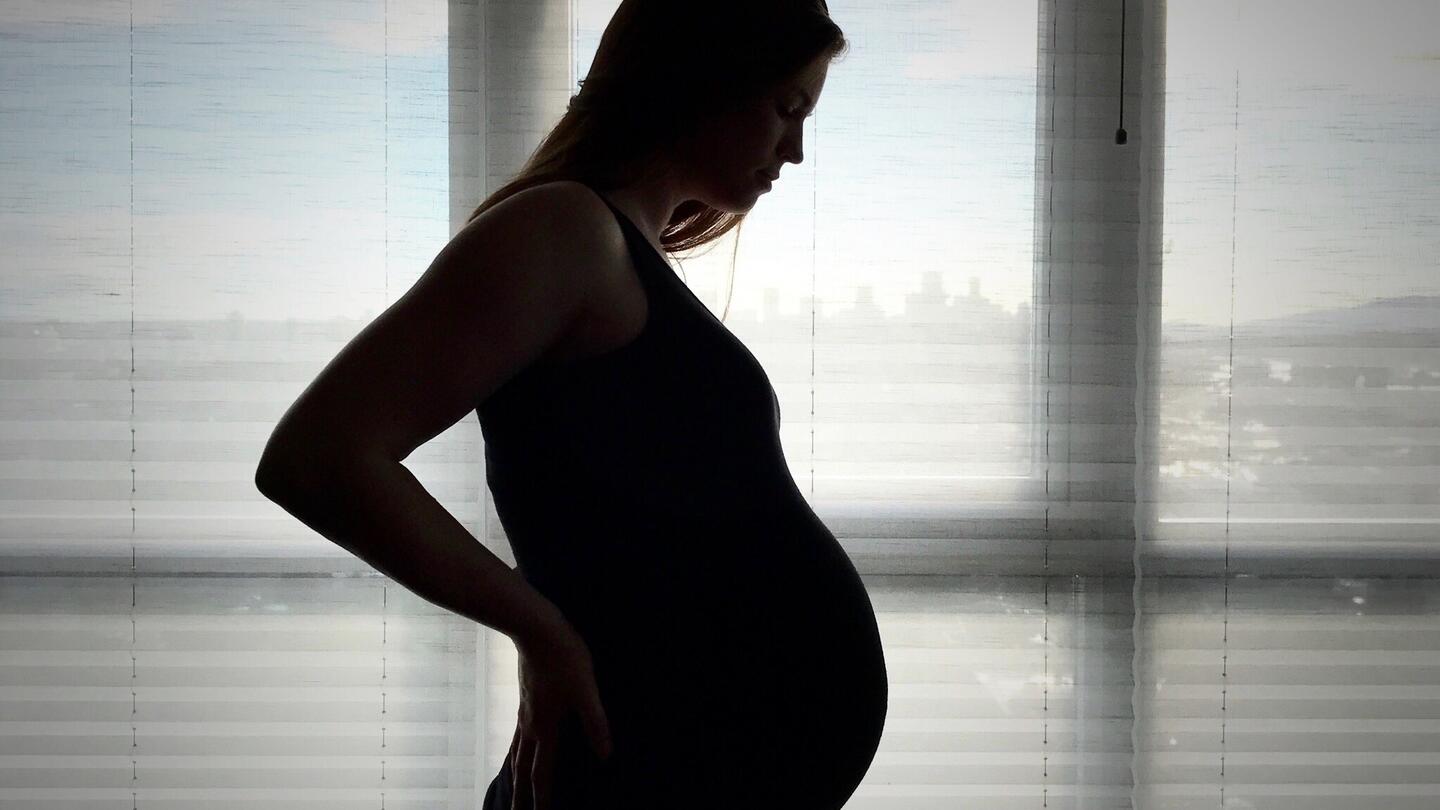 A backlit, pregnant woman stands in front of a window