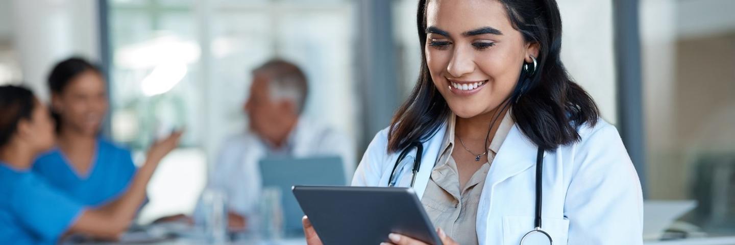 A woman is seated, wearing a white lab coat and stethoscope smiles as she looks at the screen of a tablet
