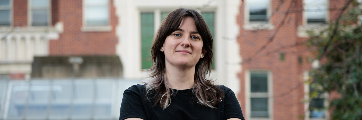 A woman stands in front a building with her arms crossed and a slight smile