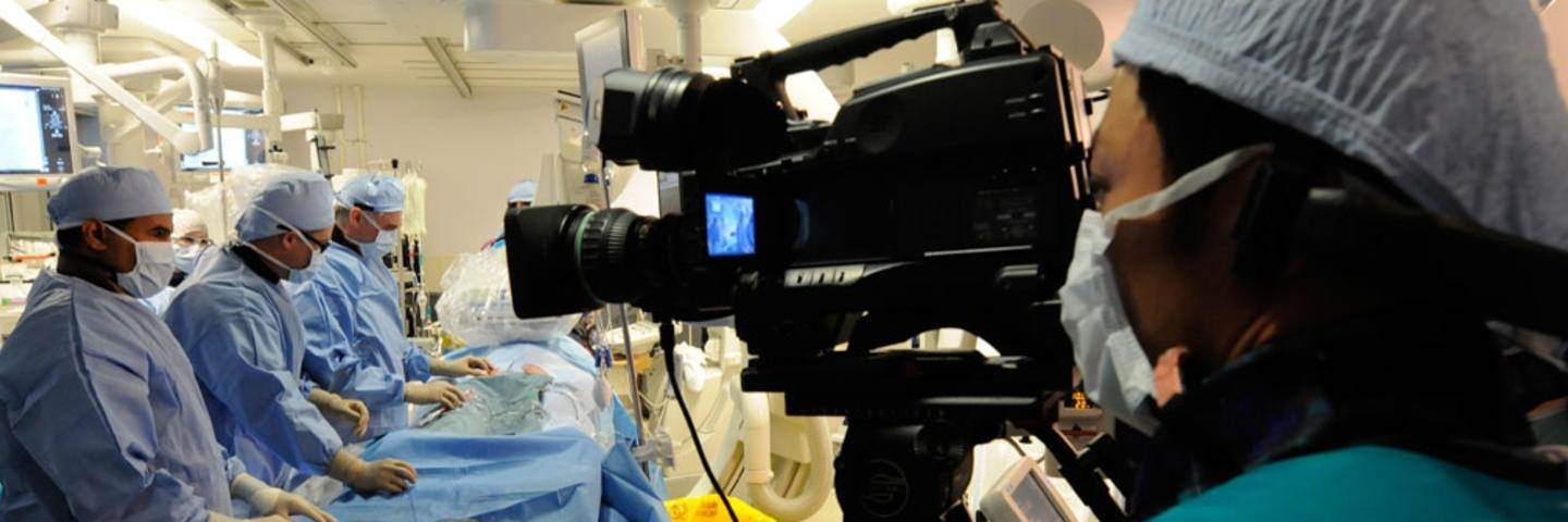 Three people in scrubs stand over an operating table while a cameraman records
