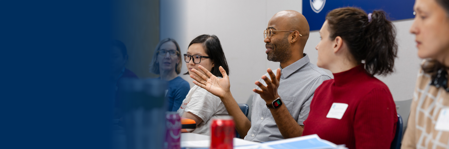 Six people sit around a table and look at a man speaking