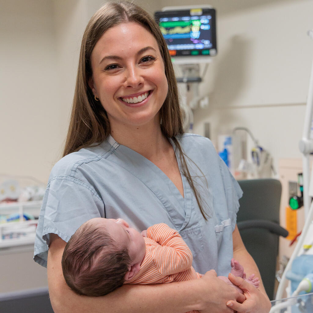 A smiling female nurse wearing blue scrubs holds a newborn baby in her arms
